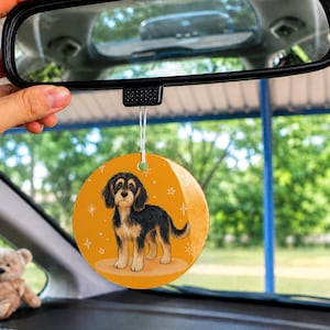 May include: A round car air freshener hanging from a rearview mirror, featuring a graphic illustration of a black and tan puppy on a solid orange background with small white star accents, suspended by a thin white string.