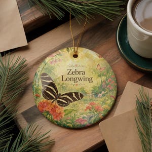 May include: Round ceramic ornament featuring a Zebra Longwing butterfly with black and white striped wings, perched on orange flowers. The ornament has a gold string for hanging and includes the text "Zebra Longwing". The background depicts a garden scene.