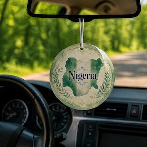 May include: A circular car air freshener hanging from a rearview mirror, featuring a green and white watercolor map of Nigeria with the word Nigeria printed in black text, surrounded by a decorative laurel wreath design on a light background.