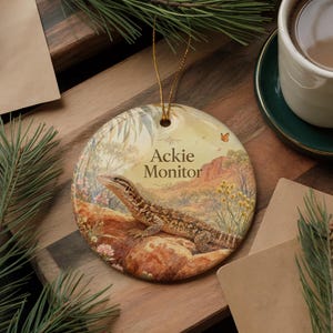 May include: A round ceramic ornament featuring an Ackie Monitor lizard in a desert landscape. The ornament has a gold string for hanging. The text "Ackie Monitor" is printed on the ornament.