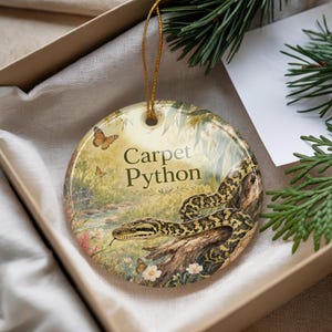 May include: Round ceramic ornament featuring a detailed illustration of a carpet python coiled on a tree branch. The ornament has a gold string for hanging and includes the text "Carpet Python". Butterflies and flowers are also depicted.