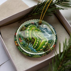 May include: A round ceramic ornament featuring a vibrant green snake coiled on a mossy branch. The background depicts a lush tropical rainforest with misty mountains. The ornament has a decorative border and a gold hanging string attached at the top.