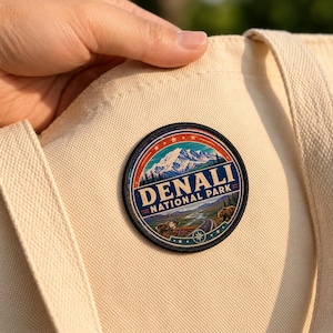 May include: A circular embroidered patch featuring a landscape of Denali National Park with a snow-capped mountain, a winding road, a caribou, and a bear. The text reads DENALI NATIONAL PARK in bold letters. The patch is attached to a cream canvas tote bag.