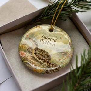 May include: A round ceramic ornament featuring a spotted python illustration. The ornament has a gold string for hanging and the text "Spotted Python". The background depicts a natural scene with flowers and butterflies.