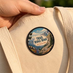 May include: A circular embroidered patch featuring a scenic mountain landscape with a winding road, pine trees, and colorful wildflowers. The text reads TRAIL RIDGE ROAD and All-American Road in dark blue and red script on a cream canvas tote bag.