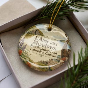 May include: Round ceramic Christmas ornament featuring a scenic illustration of L'Anse aux Meadows, Newfoundland and Labrador, Canada. The ornament has a gold string for hanging and is presented in a gift box with a sprig of evergreen.