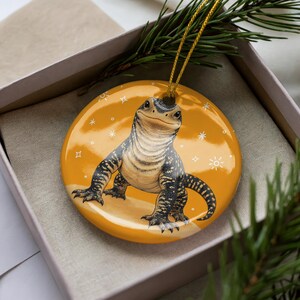 May include: A round, glossy, mustard-yellow ceramic ornament featuring a detailed illustration of a black and white lizard with a textured, scaly appearance. The lizard is centered on the ornament, surrounded by small, white, hand-drawn star and sun icons.