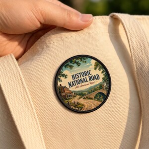 May include: A circular embroidered patch featuring a landscape illustration of a stone bridge and a building along a winding road. The text reads HISTORIC NATIONAL ROAD All-American Road. The patch is attached to a cream-colored canvas tote bag.