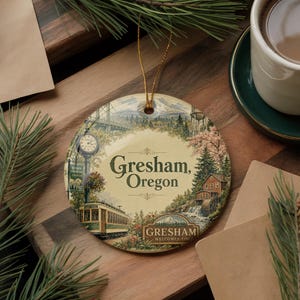 May include: A round ceramic ornament featuring a vintage illustration of Gresham, Oregon. The design includes a bridge, clock tower, streetcar, and the text "Gresham, Oregon". A gold string is attached for hanging.