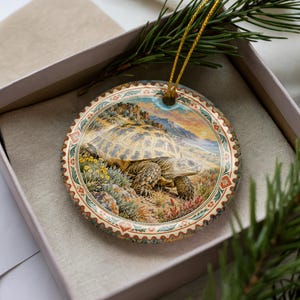 May include: A round ceramic ornament featuring a detailed illustration of a tortoise in a desert landscape with rocky terrain and yellow flowers. The ornament has a decorative border with floral patterns and includes a gold hanging string.