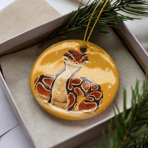 May include: A round ceramic ornament featuring a brown and tan patterned snake illustration on a mustard yellow background with white star and sun motifs. The ornament has a glossy finish and includes a gold hanging cord attached at the top.