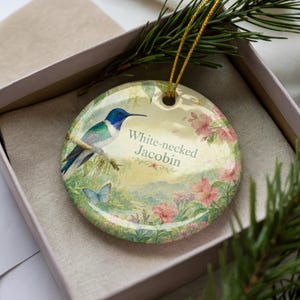 May include: Round ceramic ornament featuring a White-necked Jacobin hummingbird perched on a branch. The ornament has a gold string for hanging and is decorated with pink flowers and a blue butterfly. The text "White-necked Jacobin" is printed on the ornament.