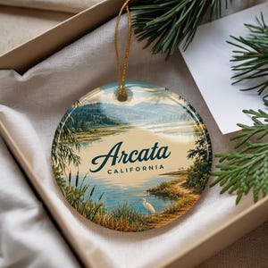 May include: A round ceramic ornament featuring a landscape illustration of a lake with a white heron in the foreground, hills in the distance, and the text Arcata CALIFORNIA printed in dark blue script and block lettering with a gold hanging cord.