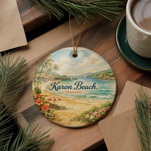 May include: Round ceramic ornament featuring a beach scene with the text "Karon Beach, Thailand." The ornament depicts a beach with palm trees, a pier, and a red and white umbrella. A gold string is attached for hanging.