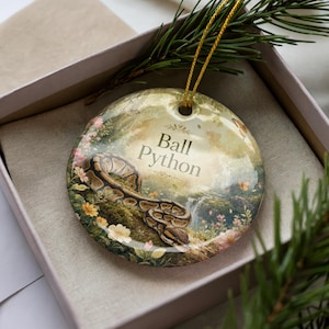 May include: A round ceramic ornament featuring a ball python in a naturalistic setting with the text "Ball Python". The ornament has a gold string for hanging and is presented in a gift box with greenery.
