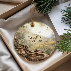 May include: A round ceramic ornament featuring a detailed illustration of a Bredl's Python snake on a log, set against a scenic background. The ornament has a gold string for hanging and is presented in a gift box.