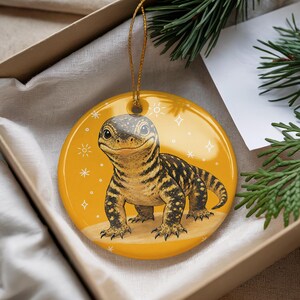 May include: A round, glossy, yellow ceramic ornament featuring a detailed illustration of a lizard with black and yellow patterned skin. The lizard is centered on the ornament, which is decorated with small white star and sun icons and has a gold string.