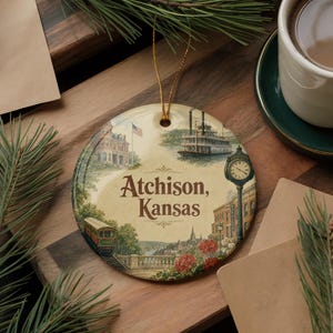 May include: Round ceramic Christmas ornament featuring a vintage illustration of Atchison, Kansas. The design includes a steamboat, a train, a clock tower, and the text "Atchison, Kansas". The ornament has a gold string for hanging.