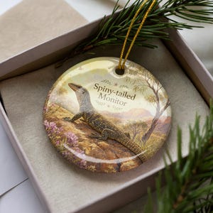 May include: A round ceramic ornament featuring a Spiny-tailed Monitor lizard illustration. The ornament has a gold string for hanging and is presented in a gift box. The lizard is depicted on a landscape background with the text "Spiny-tailed Monitor".
