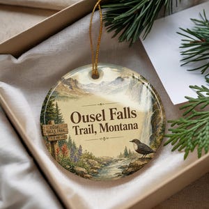 May include: Round ceramic ornament featuring a scenic landscape with a waterfall, river, and mountains. The text "Ousel Falls Trail, Montana" is displayed. A small bird is perched on a rock. The ornament is in a gift box.