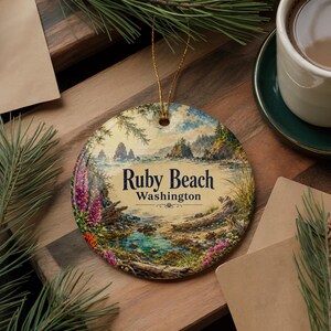 May include: A round ceramic ornament featuring a scenic illustration of Ruby Beach, Washington, with coastal rock formations, tide pools, and wildflowers. The text Ruby Beach Washington is printed in the center. A gold string is attached for hanging.