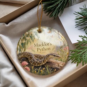 May include: Round ceramic ornament featuring a detailed illustration of a Macklot's Python snake resting on a log, surrounded by flowers and foliage. The ornament has a gold string for hanging and the text "Macklot's Python".