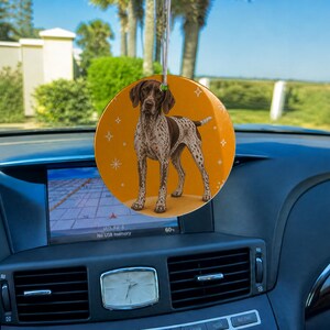May include: A round, hanging car ornament featuring a brown and white spotted German Shorthaired Pointer dog illustration against a solid orange background with small white star accents, suspended by a thin silver string from a rearview mirror.