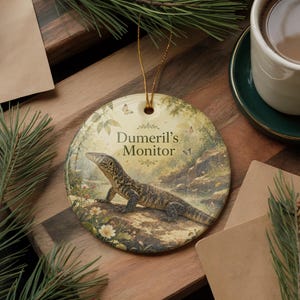 May include: Round ceramic ornament featuring a detailed illustration of a Dumeril's Monitor lizard in a natural setting. The ornament has a gold string for hanging and the text "Dumeril's Monitor". The background includes foliage, water, and butterflies.