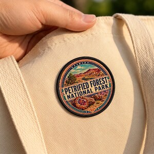 May include: A circular embroidered patch featuring a landscape of Petrified Forest National Park with red rock formations, a winding road, and a cross-section of petrified wood, attached to a cream-colored canvas tote bag with visible fabric texture.