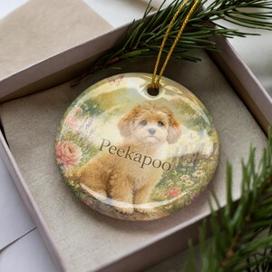 May include: A round ceramic ornament featuring a Peekapoo dog illustration in a garden setting with pink roses and butterflies. The ornament has a gold string for hanging and is presented in a gift box.