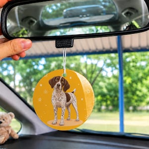 May include: A circular car air freshener hanging from a rearview mirror, featuring a brown and white spotted dog illustration on a solid mustard yellow background with small white star and paw print accents printed on the surface.