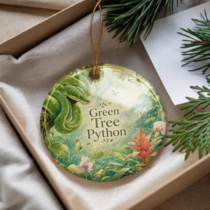 May include: A round ceramic ornament featuring a green tree python in a jungle scene. The ornament has the text "Green Tree Python" and is accented with butterflies and flowers. It is displayed in a gift box.