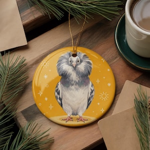 May include: A round ceramic ornament featuring a central illustration of a grey Jacobin pigeon with a large, fluffy neck ruff. The background is a solid mustard yellow color decorated with small white line-drawn suns and four-pointed stars.