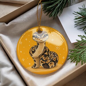 May include: A round, glossy ceramic ornament featuring a cartoon-style ball python with brown and black markings. The snake is centered on a vibrant yellow background decorated with small white star and sun illustrations. A gold string is attached.