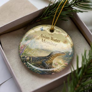 May include: Round ceramic ornament featuring an Asian Water Monitor lizard in a naturalistic setting with butterflies. The ornament has a gold string for hanging and is presented in a gift box.