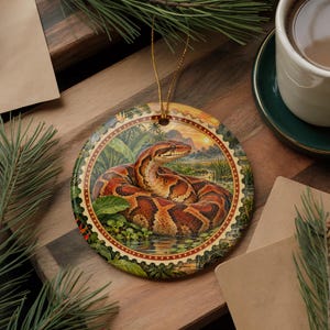 May include: A round ceramic ornament featuring a detailed illustration of a coiled brown and orange snake in a tropical swamp setting. The artwork is framed by a decorative scalloped border with a gold-toned string loop for hanging purposes.
