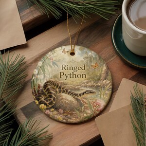 A round ornament featuring a snake and flowers, with the text "Ringed Python" displayed on it, is placed on a wooden surface next to a cup of coffee and some pine branches.