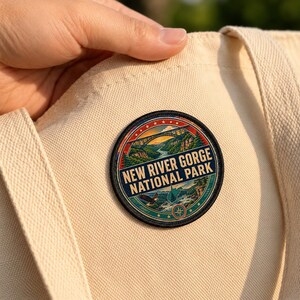 May include: A circular embroidered patch featuring a landscape of the New River Gorge National Park with a bridge, river, and eagle. The text NEW RIVER GORGE NATIONAL PARK is printed in bold letters. The patch is attached to a cream-colored canvas tote bag.