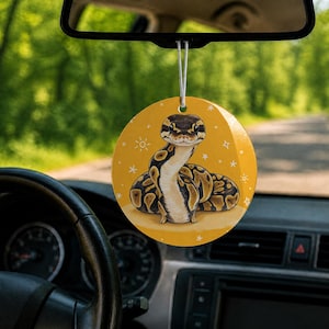 May include: A round, yellow car air freshener hanging from a rearview mirror. The ornament features a detailed illustration of a brown and black ball python snake with a light-colored underbelly, surrounded by small white star and sun icons.