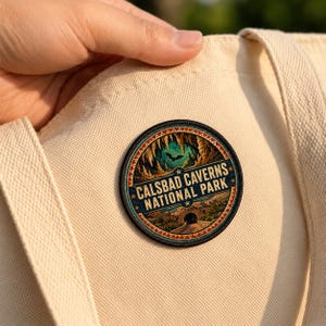 May include: A circular embroidered patch featuring a landscape of Carlsbad Caverns National Park, including stalactites, a flying bat, and a stone entrance arch, with the text "CALSBAD CAVERNS NATIONAL PARK" attached to a beige canvas tote bag.