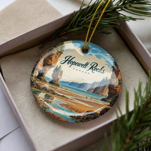May include: A round ceramic ornament featuring a printed landscape of the Hopewell Rocks in Canada. The image displays tall rock formations, a sandy beach, and blue water under a sky. The text Hopewell Rocks CANADA is printed in dark blue.