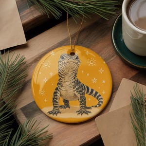 May include: A round ceramic ornament featuring a detailed illustration of a lizard with black and yellow patterned skin. The background is a solid mustard yellow color decorated with white line drawings of small suns and stars, complete with a gold string.