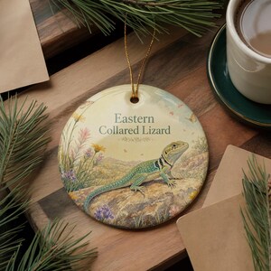 May include: A round ceramic ornament featuring an Eastern Collared Lizard illustration. The lizard is depicted in shades of green and blue, resting on a rock. The ornament has a gold string for hanging and includes the text "Eastern Collared Lizard".