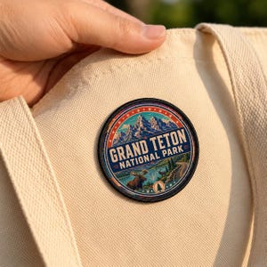 May include: A circular embroidered patch featuring a landscape of Grand Teton National Park with snow-capped mountains, a lake, a moose, and a winding road. The text reads GRAND TETON NATIONAL PARK. The patch is attached to a cream-colored canvas tote bag.