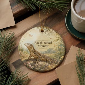 May include: A round ceramic ornament featuring a Rough-necked Monitor lizard illustration. The lizard is depicted in a naturalistic setting with butterflies and foliage. The ornament has a gold string for hanging.