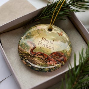 May include: A round ceramic ornament featuring a detailed illustration of a blood python in a natural setting. The ornament has a gold string for hanging and the words "Blood Python" are printed on the front.