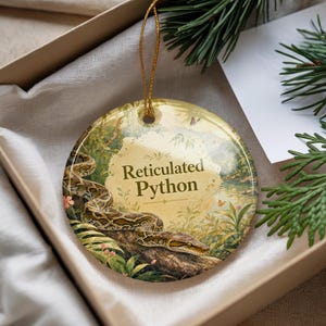 May include: A round ceramic ornament featuring a reticulated python design. The ornament has a gold string for hanging. The background shows a jungle scene with the text "Reticulated Python". The ornament is in a gift box.