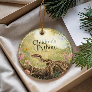 May include: A round ceramic ornament featuring a detailed illustration of a snake on a log, with the text "Children's Python". The background includes flowers, butterflies, and foliage. The ornament has a gold string for hanging.