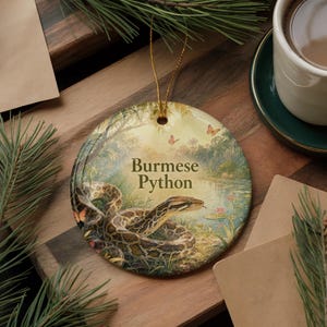 May include: A round ceramic ornament featuring a detailed illustration of a Burmese python in a natural habitat. The ornament has a gold string for hanging and the text "Burmese Python". The background includes a river, trees, and butterflies.