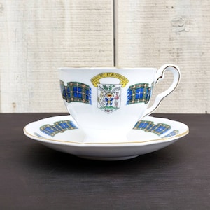 May include: White teacup and saucer set with gold trim. The cup and saucer feature a blue and green plaid pattern and a crest with the text "NIVIE HEC ET ALTERAVIRIC". A decorative tea set.